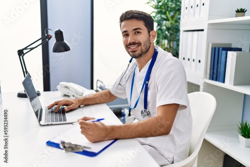 Young hispanic man wearing doctor uniform writing on document working at clinic