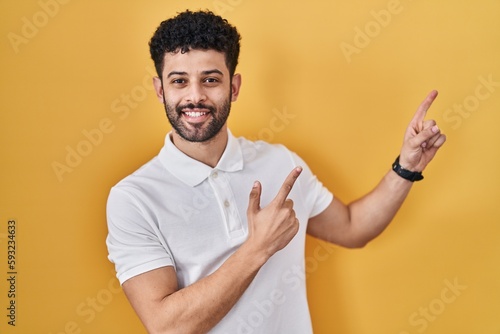 Arab man standing over yellow background smiling and looking at the camera pointing with two hands and fingers to the side.