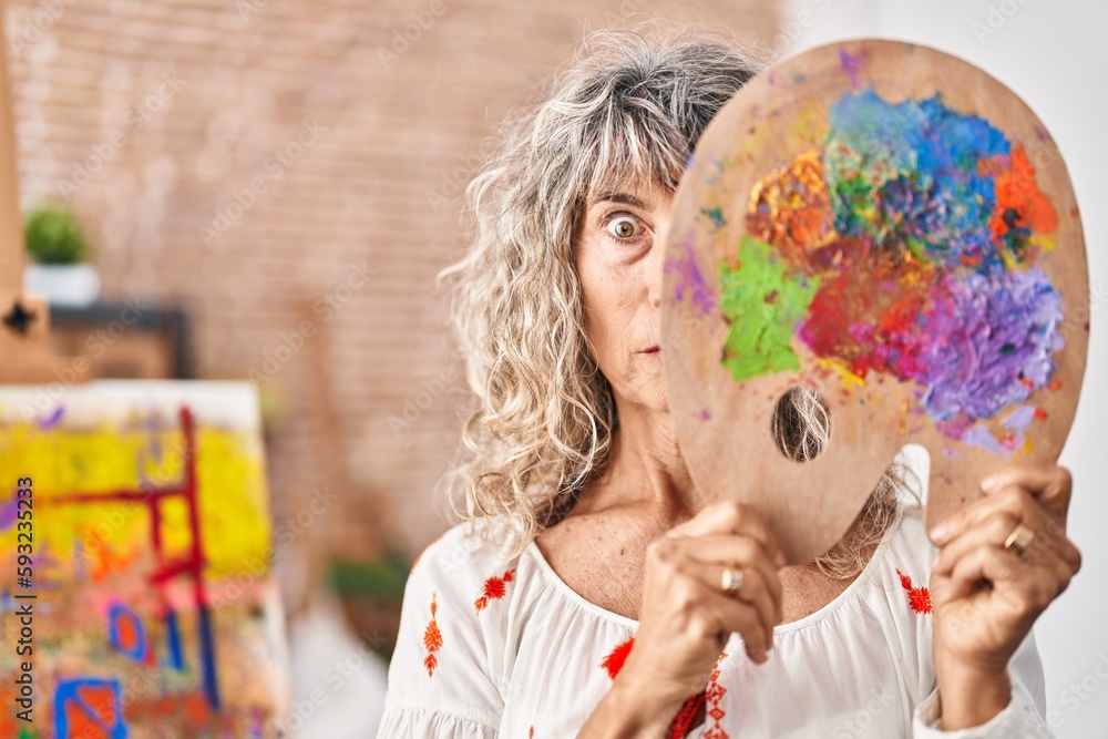 Fototapeta premium Middle age woman holding painter palette covering face clueless and confused expression. doubt concept.