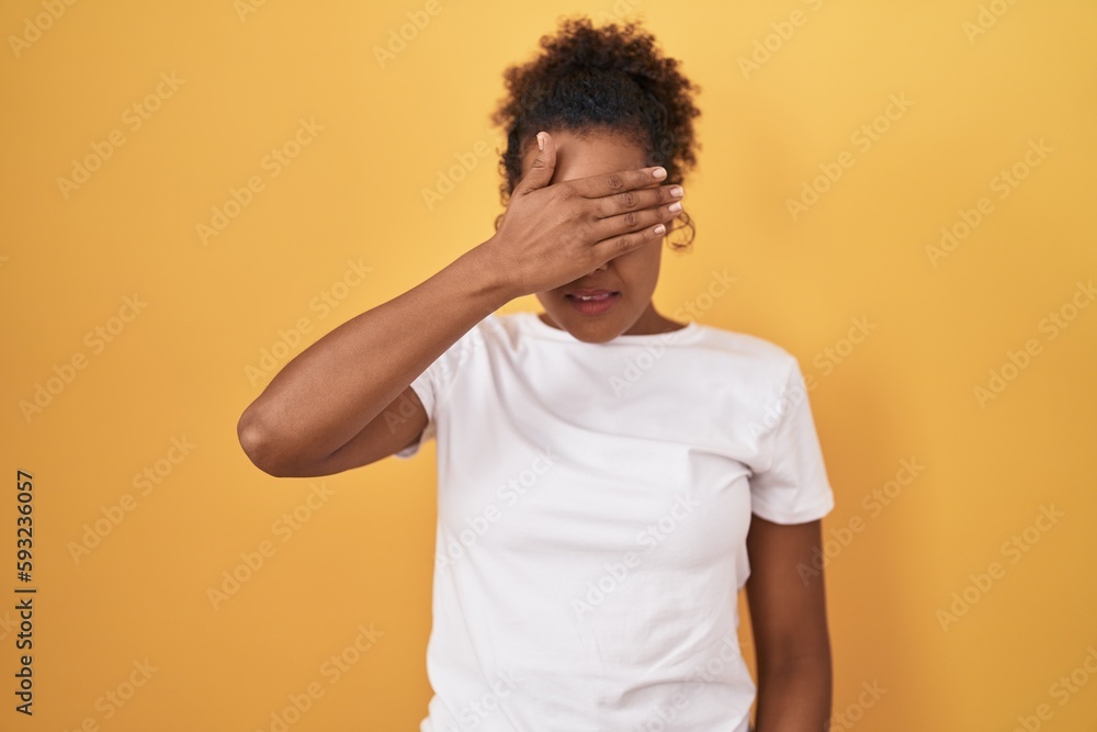 Young hispanic woman with curly hair standing over yellow background covering eyes with hand, looking serious and sad. sightless, hiding and rejection concept Young hispanic woman with curly hair standing over yellow background covering eyes with hand, looking serious and sad. sightless, hiding and rejection concept