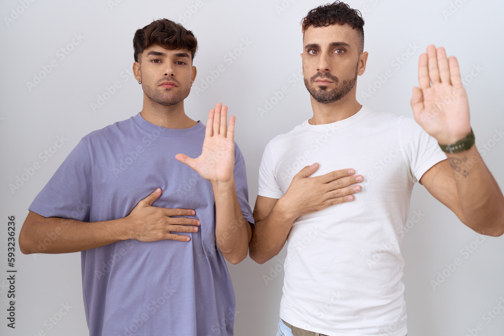 Homosexual gay couple standing over white background swearing with hand on chest and open palm, making a loyalty promise oath Homosexual gay couple standing over white background swearing with hand on chest and open palm, making a loyalty promise oath