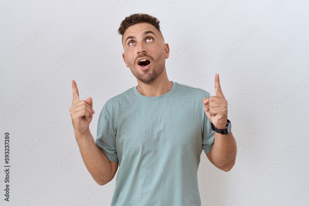 Hispanic man with beard standing over white background amazed and surprised looking up and pointing with fingers and raised arms. Hispanic man with beard standing over white background amazed and surprised looking up and pointing with fingers and raised arms.