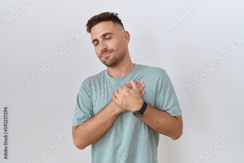 Hispanic man with beard standing over white background smiling with hands on chest with closed eyes and grateful gesture on face. health concept.