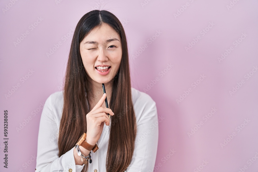 Chinese young woman holding pencil over pink background winking looking at the camera with sexy expression, cheerful and happy face.