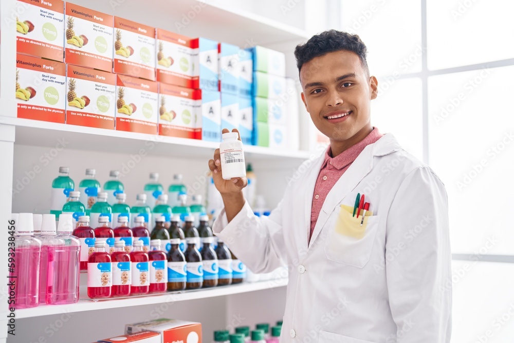 Young latin man pharmacist smiling confident holding pills bottle at pharmacy