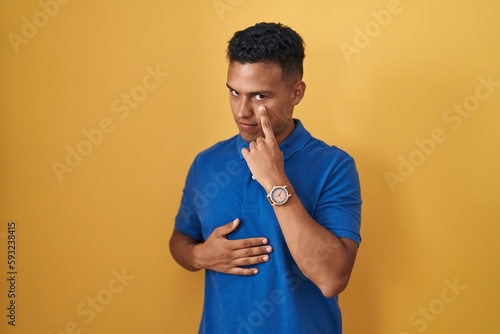 Young hispanic man standing over yellow background pointing to the eye watching you gesture, suspicious expression