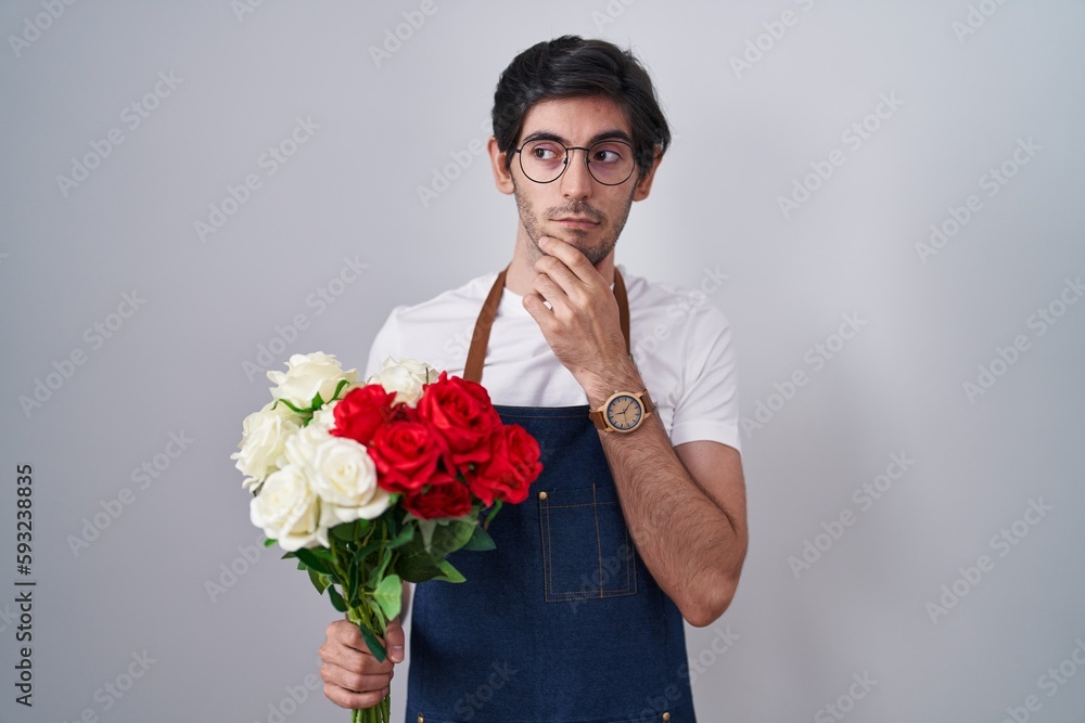Young hispanic man holding bouquet of white and red roses thinking worried about a question, concerned and nervous with hand on chin Young hispanic man holding bouquet of white and red roses thinking worried about a question, concerned and nervous with hand on chin