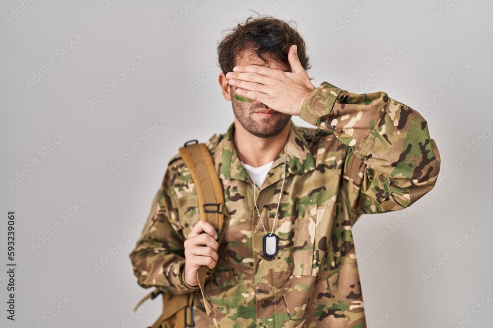 Hispanic young man wearing camouflage army uniform covering eyes with hand, looking serious and sad. sightless, hiding and rejection concept