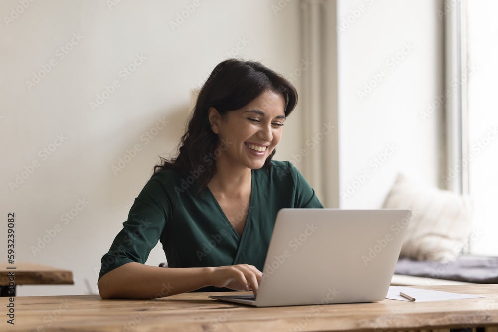 Cheerful young worker woman sit at desk with laptop at home office, look at screen, feel happy and optimistic after completing successful project, use internet resource, work on new motivation article Cheerful young worker woman sit at desk with laptop at home office, look at screen, feel happy and optimistic after completing successful project, use internet resource, work on new motivation article