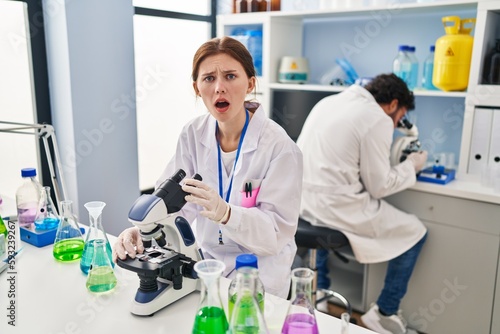 Young two people working at scientist laboratory scared and amazed with open mouth for surprise  disbelief face
