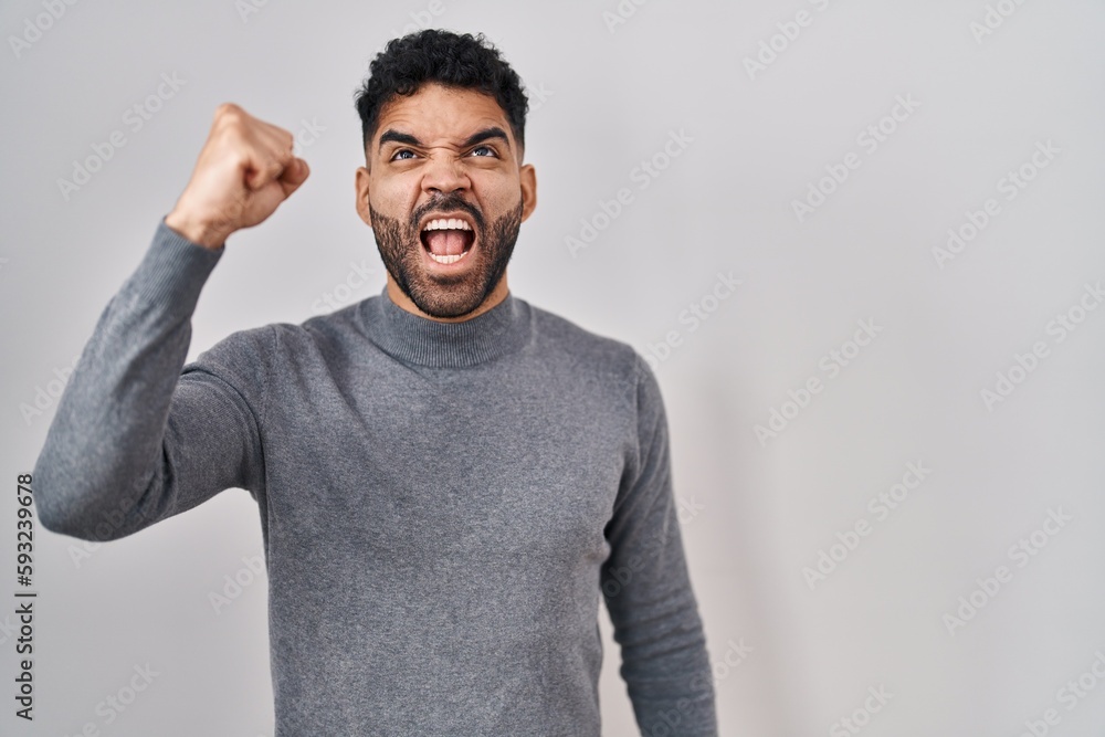 Hispanic man with beard standing over white background angry and mad raising fist frustrated and furious while shouting with anger. rage and aggressive concept. Hispanic man with beard standing over white background angry and mad raising fist frustrated and furious while shouting with anger. rage and aggressive concept.