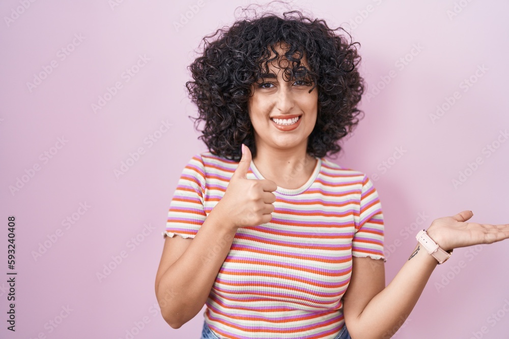 Young middle east woman standing over pink background showing palm hand and doing ok gesture with thumbs up, smiling happy and cheerful Young middle east woman standing over pink background showing palm hand and doing ok gesture with thumbs up, smiling happy and cheerful