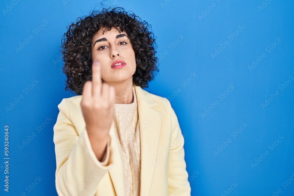 Young brunette woman with curly hair standing over blue background showing middle finger, impolite and rude fuck off expression Young brunette woman with curly hair standing over blue background showing middle finger, impolite and rude fuck off expression