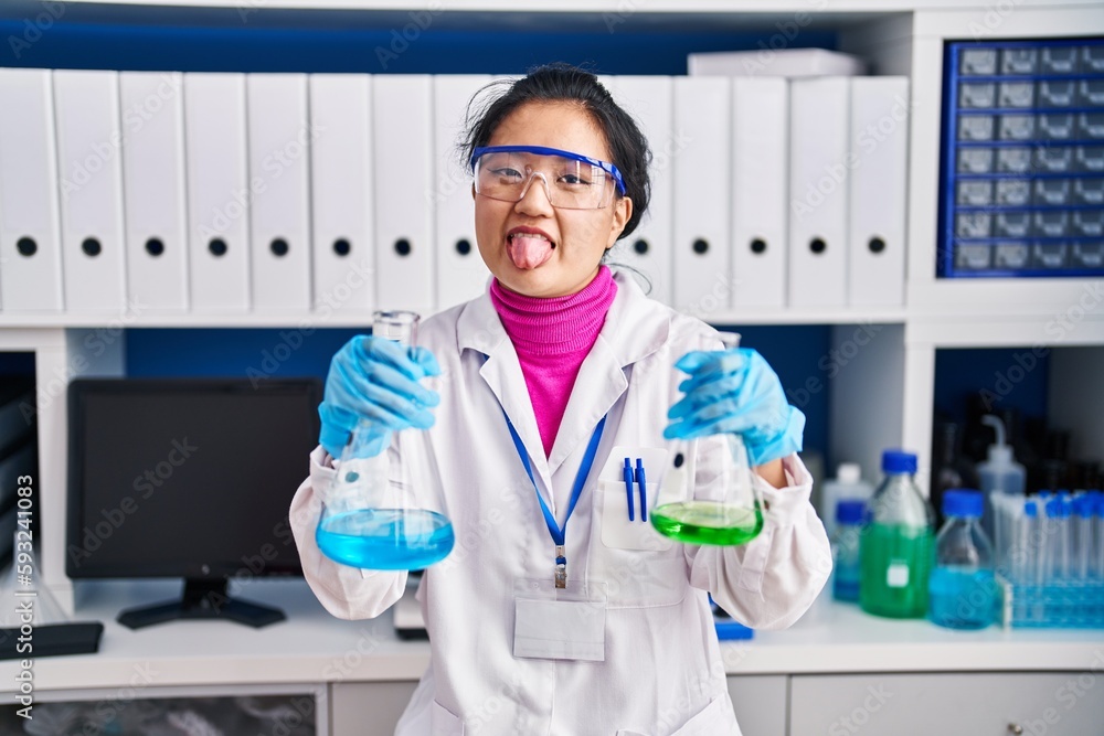 Young asian woman working at scientist laboratory sticking tongue out happy with funny expression.
