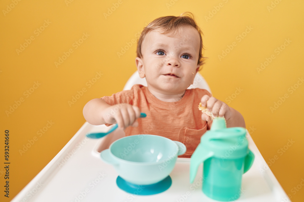 Adorable blond toddler sitting on highchair eating snack over isolated yellow background Adorable blond toddler sitting on highchair eating snack over isolated yellow background