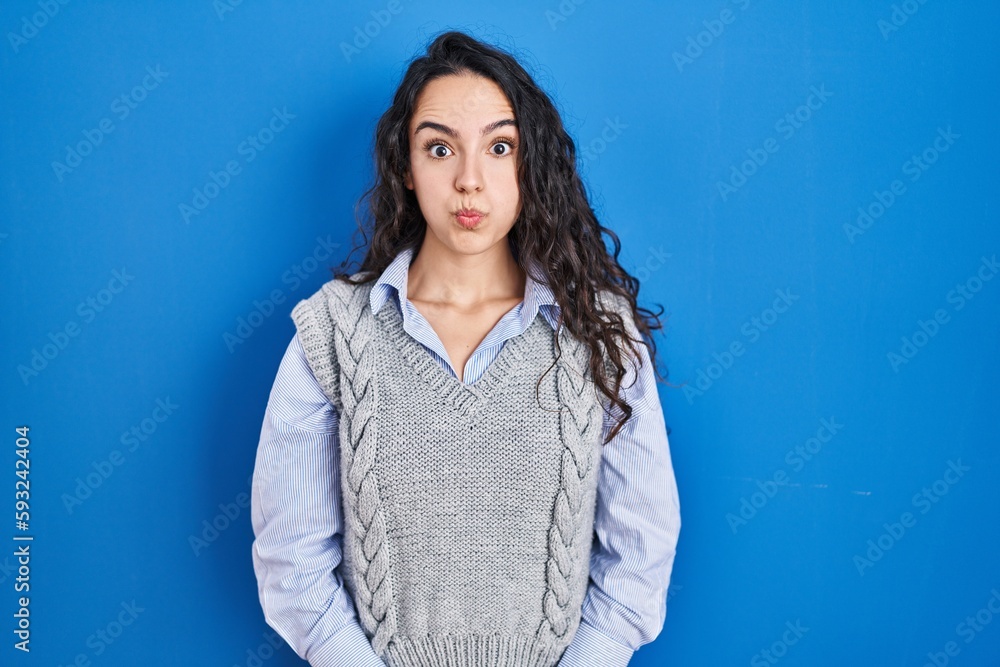 Fototapeta premium Young brunette woman standing over blue background puffing cheeks with funny face. mouth inflated with air, crazy expression.