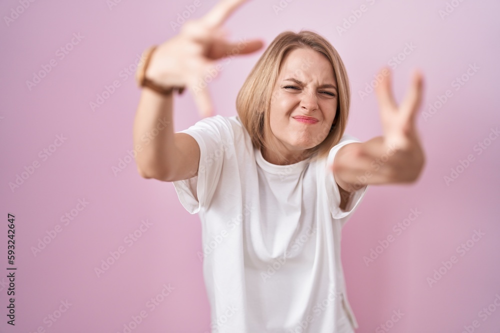 Young caucasian woman standing over pink background shouting frustrated with rage, hands trying to strangle, yelling mad
