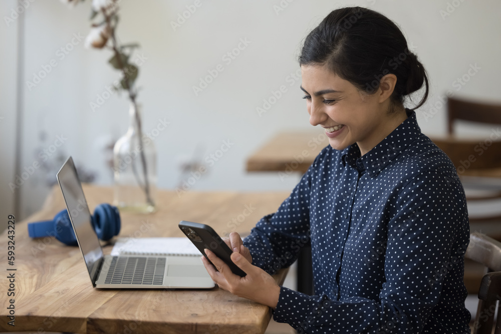 Happy Indian woman enjoy on-line chat, distracted from work or study sit at desk with laptop smile, read sms, share messages, ordering goods use easy, comfort remote electronic services at workplace Happy Indian woman enjoy on-line chat, distracted from work or study sit at desk with laptop smile, read sms, share messages, ordering goods use easy, comfort remote electronic services at workplace