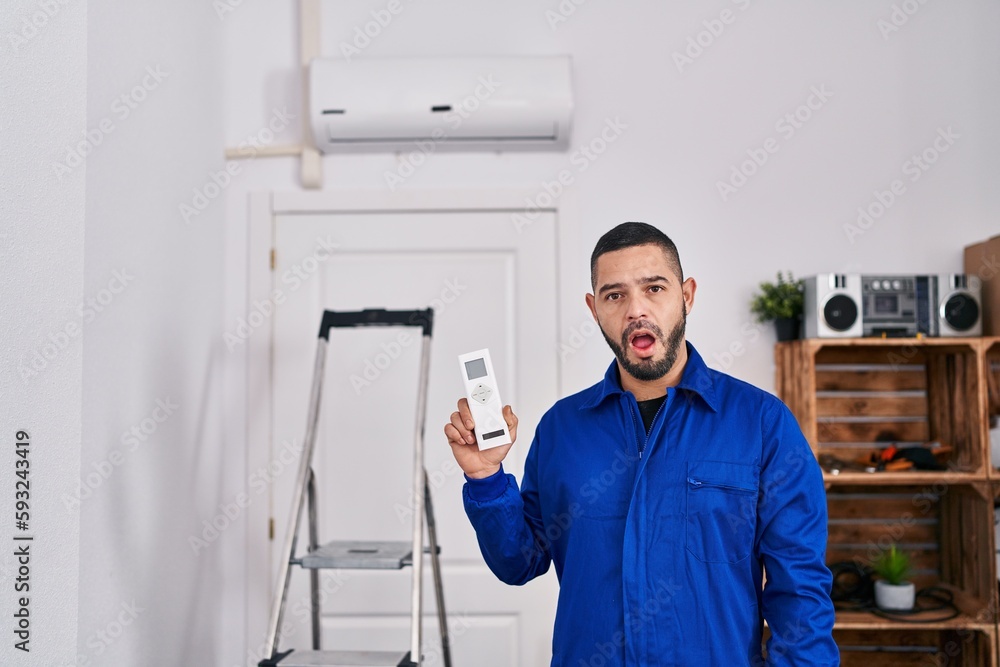 Hispanic repairman working with air conditioner scared and amazed with open mouth for surprise, disbelief face
