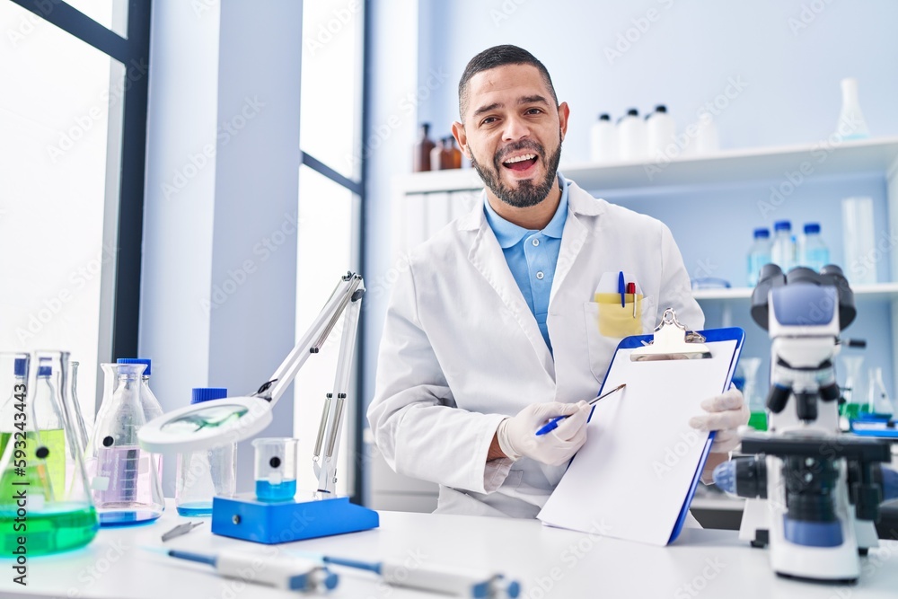 Hispanic man working at scientist laboratory holding blank clipboard celebrating crazy and amazed for success with open eyes screaming excited. Hispanic man working at scientist laboratory holding blank clipboard celebrating crazy and amazed for success with open eyes screaming excited.