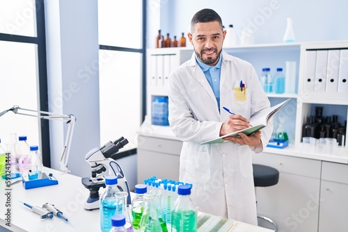 Young latin man scientist smiling confident writing on notebook at laboratory
