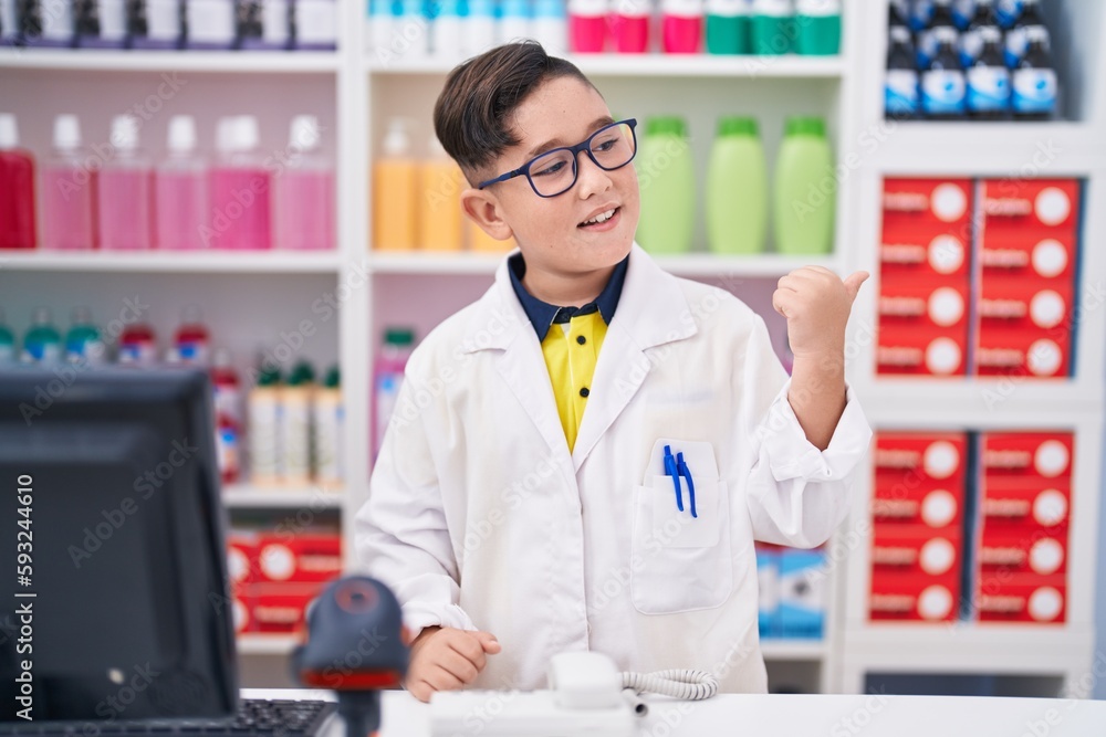 Young hispanic kid working at pharmacy drugstore pointing thumb up to the side smiling happy with open mouth Young hispanic kid working at pharmacy drugstore pointing thumb up to the side smiling happy with open mouth