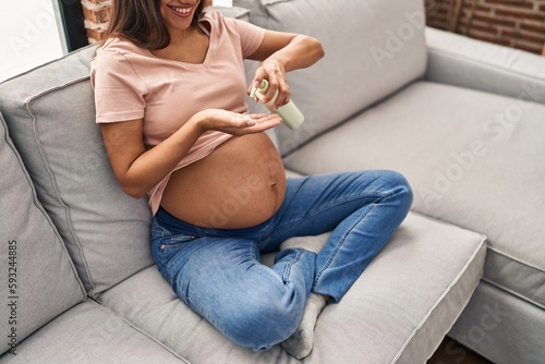 Young latin woman pregnant applying cream lotion on skin belly at home photo