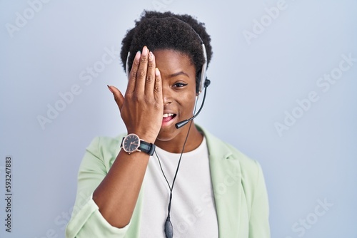 African american woman wearing call center agent headset covering one eye with hand, confident smile on face and surprise emotion.