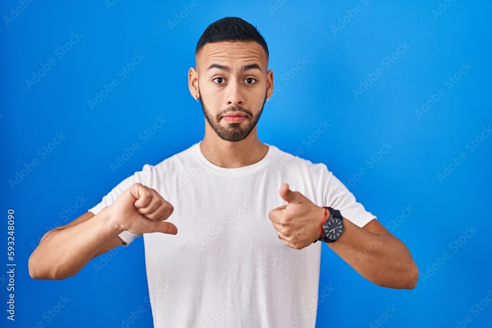 Young hispanic man standing over blue background doing thumbs up and down, disagreement and agreement expression. crazy conflict Young hispanic man standing over blue background doing thumbs up and down, disagreement and agreement expression. crazy conflict