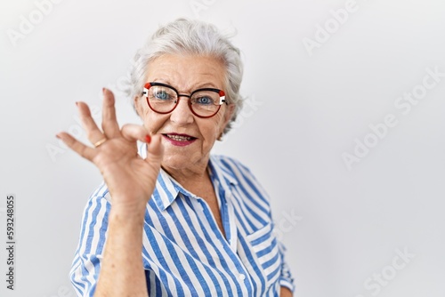 Senior woman with grey hair standing over white background smiling positive doing ok sign with hand and fingers. successful expression.