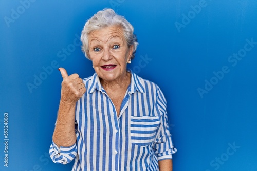 Senior woman with grey hair standing over blue background doing happy thumbs up gesture with hand. approving expression looking at the camera showing success.
