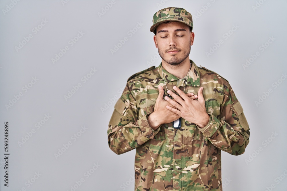 Young hispanic man wearing camouflage army uniform smiling with hands on chest with closed eyes and grateful gesture on face. health concept. Young hispanic man wearing camouflage army uniform smiling with hands on chest with closed eyes and grateful gesture on face. health concept.