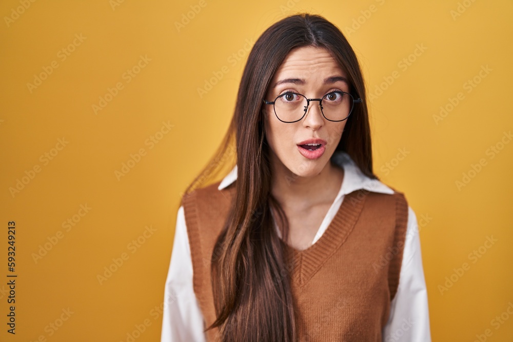 Young brunette woman standing over yellow background wearing glasses afraid and shocked with surprise expression, fear and excited face. Young brunette woman standing over yellow background wearing glasses afraid and shocked with surprise expression, fear and excited face.