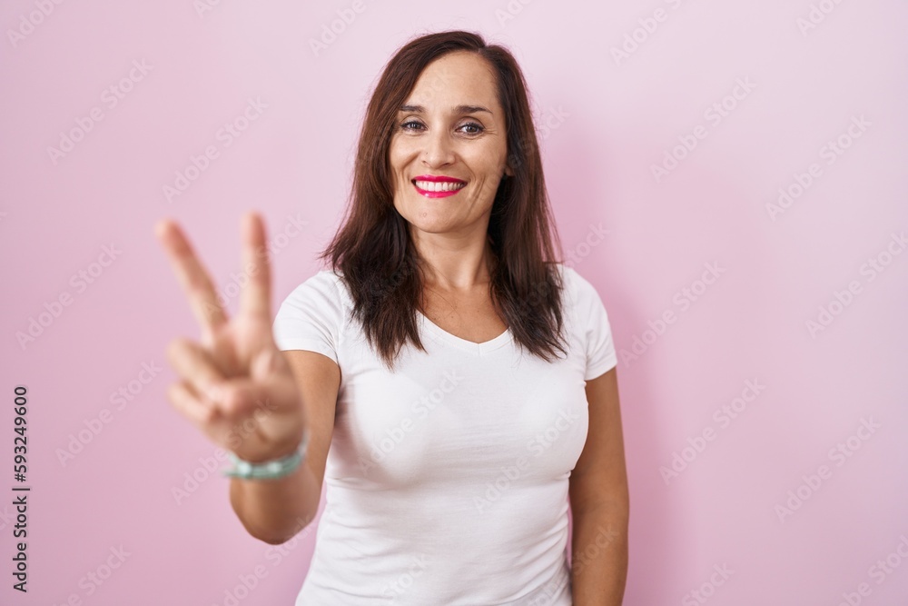 Middle age brunette woman standing over pink background smiling looking to the camera showing fingers doing victory sign. number two. Middle age brunette woman standing over pink background smiling looking to the camera showing fingers doing victory sign. number two.