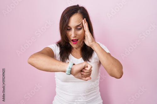 Middle age brunette woman standing over pink background looking at the watch time worried, afraid of getting late