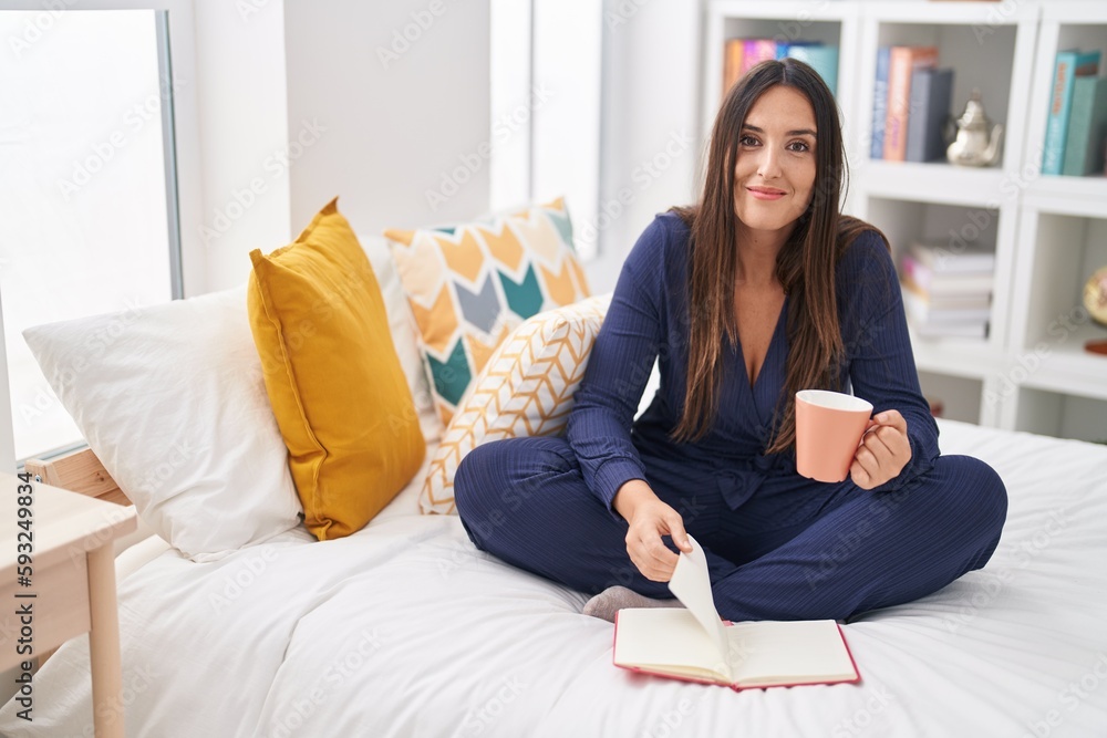 Young beautiful hispanic woman drinking cup of coffee reading book at bedroom Young beautiful hispanic woman drinking cup of coffee reading book at bedroom