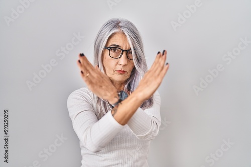 Middle age woman with grey hair standing over white background rejection expression crossing arms doing negative sign  angry face