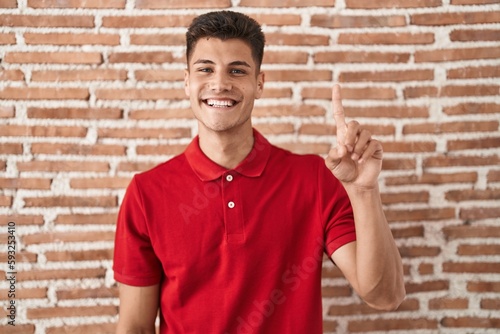 Young hispanic man standing over bricks wall showing and pointing up with finger number one while smiling confident and happy.
