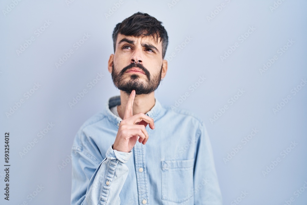 Young hispanic man with beard standing over blue background thinking concentrated about doubt with finger on chin and looking up wondering Young hispanic man with beard standing over blue background thinking concentrated about doubt with finger on chin and looking up wondering