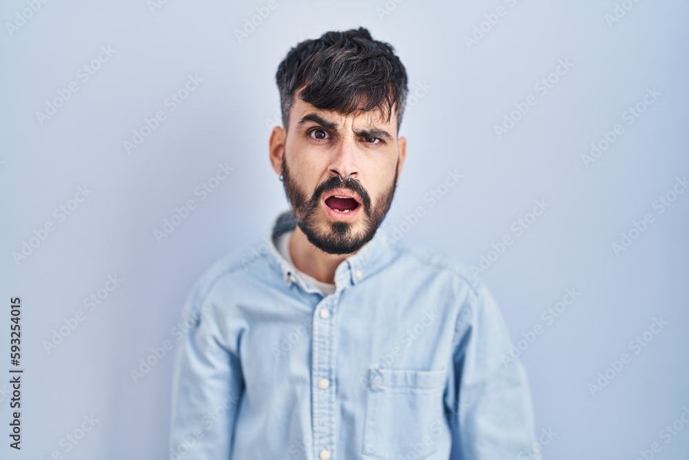 Young hispanic man with beard standing over blue background in shock face, looking skeptical and sarcastic, surprised with open mouth Young hispanic man with beard standing over blue background in shock face, looking skeptical and sarcastic, surprised with open mouth