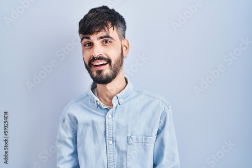 Young hispanic man with beard standing over blue background smiling cheerful with open arms as friendly welcome, positive and confident greetings
