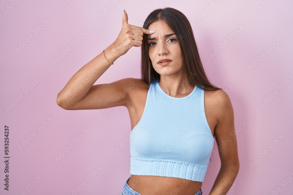 Young brunette woman standing over pink background pointing unhappy to pimple on forehead, ugly infection of blackhead. acne and skin problem