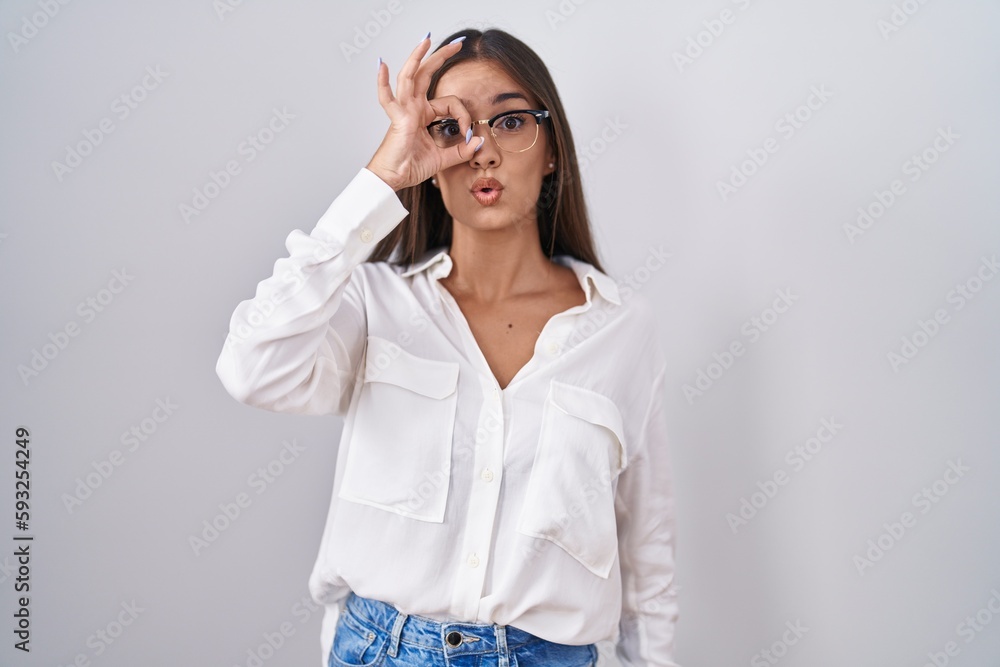 Young brunette woman wearing glasses doing ok gesture shocked with surprised face, eye looking through fingers. unbelieving expression. Young brunette woman wearing glasses doing ok gesture shocked with surprised face, eye looking through fingers. unbelieving expression.