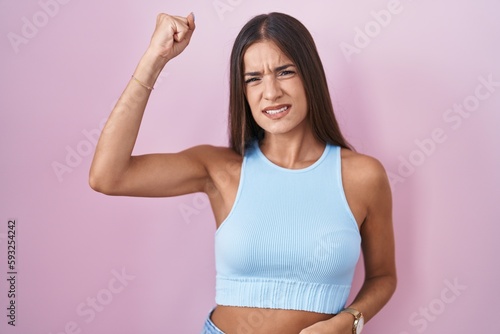 Young brunette woman standing over pink background angry and mad raising fist frustrated and furious while shouting with anger. rage and aggressive concept. © Krakenimages.com