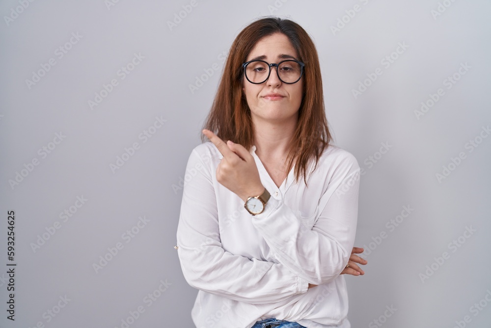 Brunette woman standing over white isolated background pointing with hand finger to the side showing advertisement, serious and calm face