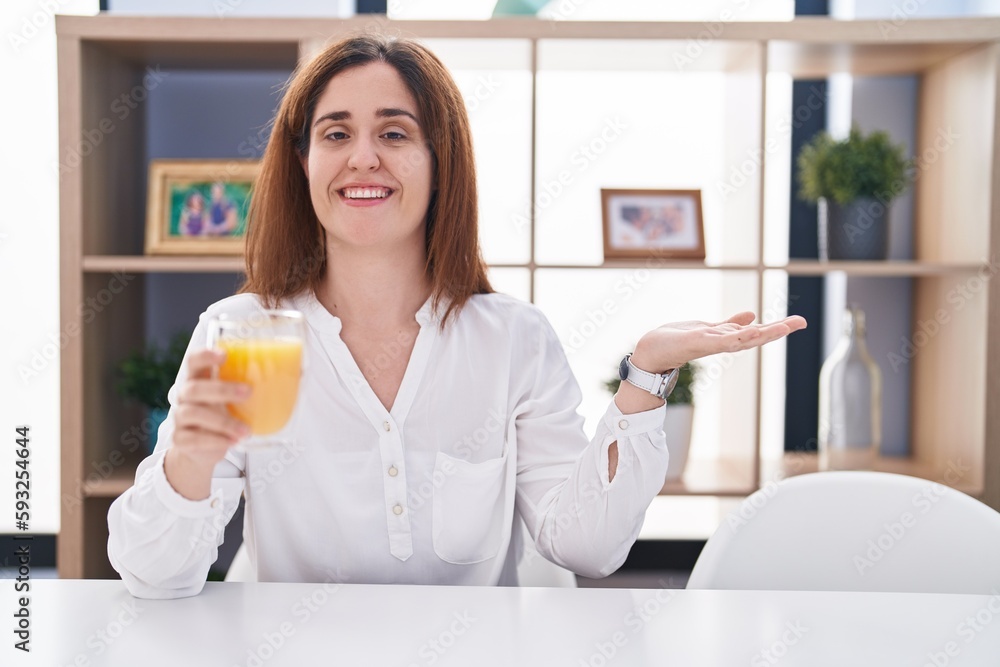 Brunette woman drinking glass of orange juice smiling cheerful presenting and pointing with palm of hand looking at the camera. Brunette woman drinking glass of orange juice smiling cheerful presenting and pointing with palm of hand looking at the camera.