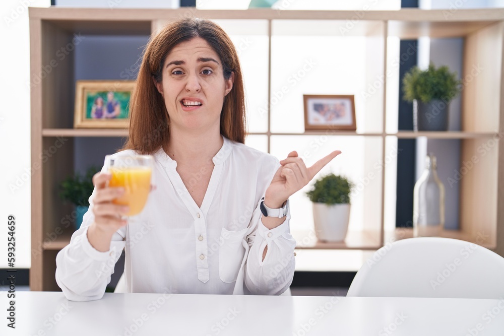 Brunette woman drinking glass of orange juice pointing aside worried and nervous with forefinger, concerned and surprised expression Brunette woman drinking glass of orange juice pointing aside worried and nervous with forefinger, concerned and surprised expression