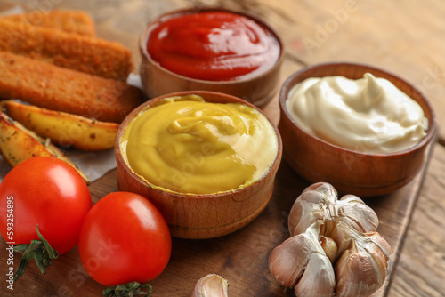 Bowls of different sauces, fish nuggets and vegetables on wooden table, closeup