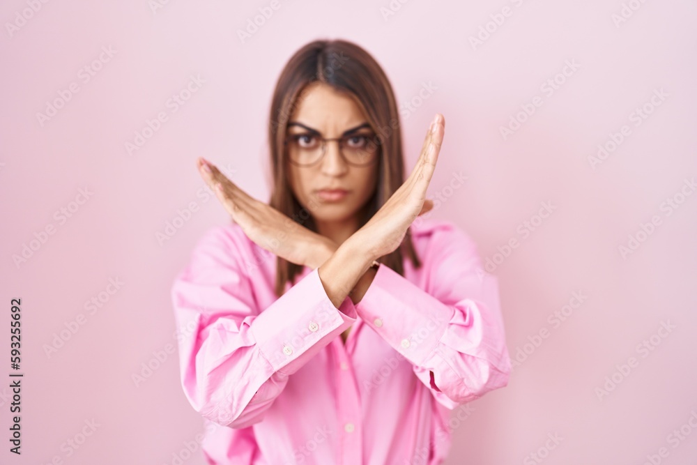 Young hispanic woman wearing glasses standing over pink background rejection expression crossing arms doing negative sign, angry face Young hispanic woman wearing glasses standing over pink background rejection expression crossing arms doing negative sign, angry face