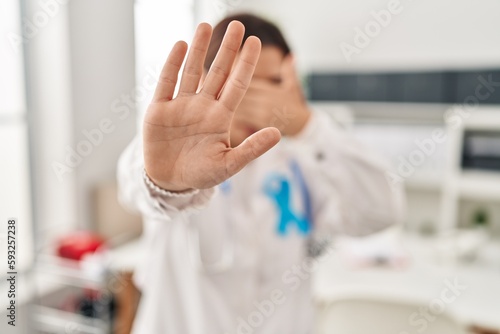 Young brunette doctor woman wearing stethoscope at the clinic covering eyes with hands and doing stop gesture with sad and fear expression. embarrassed and negative concept.